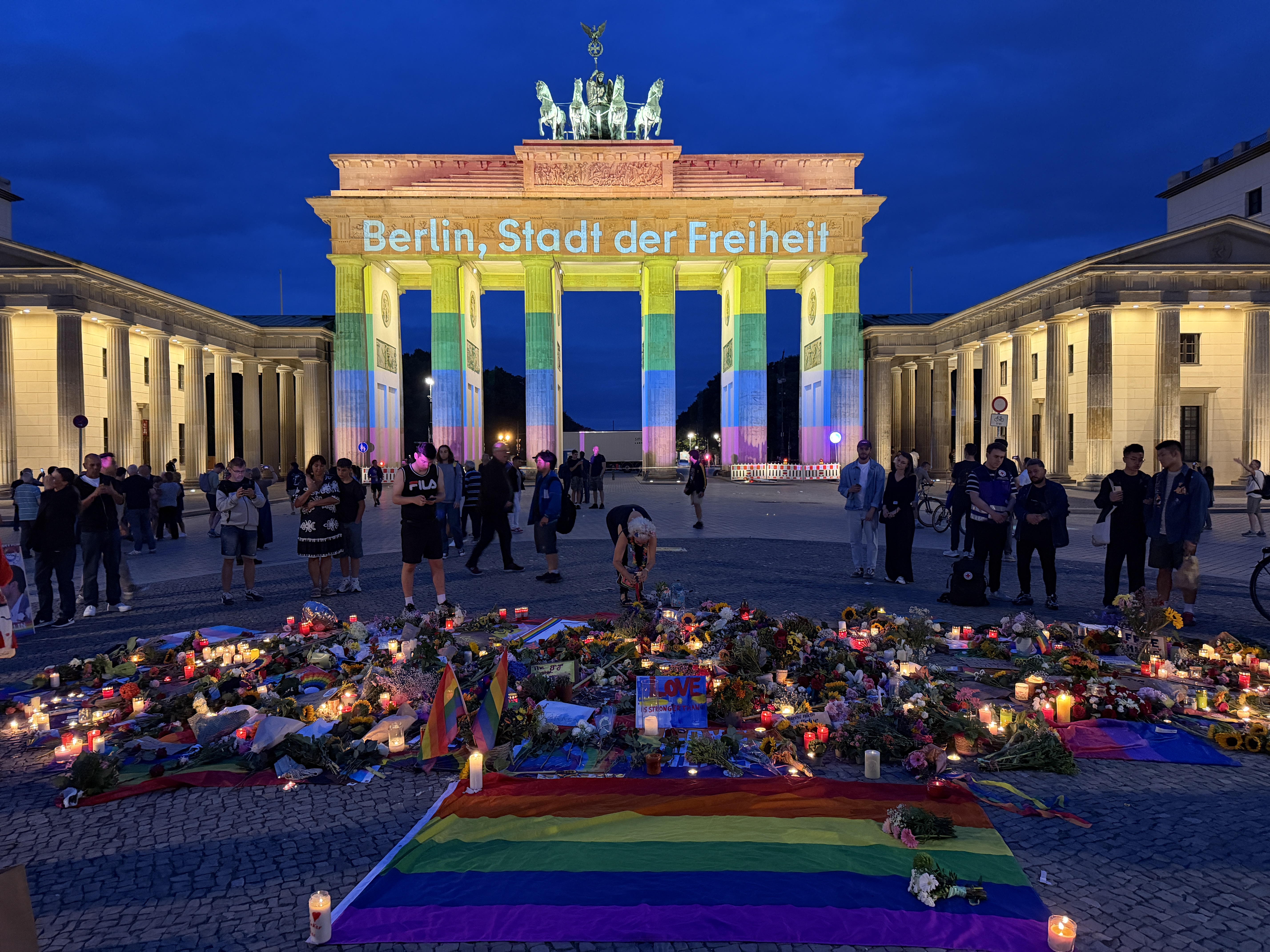 The Brandenburg Gate lit in rainbow colours with a candlelit memorial at its base, vigil of 26 July 2026